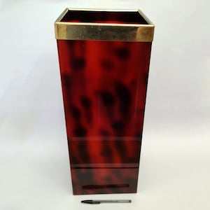 May include: A tall, rectangular wastebasket with a tortoiseshell pattern in shades of red and black. The top edge is trimmed with a gold-colored metal band. A black pen rests at the base of the wastebasket.