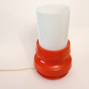 Pu&ograve; includere: Una lampada da tavolo rossa e bianca con un paralume bianco cilindrico e una base rossa con un design testurizzato.