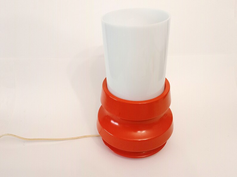Pu&ograve; includere: Una lampada da tavolo rossa e bianca con un paralume bianco cilindrico e una base rossa con un design testurizzato.