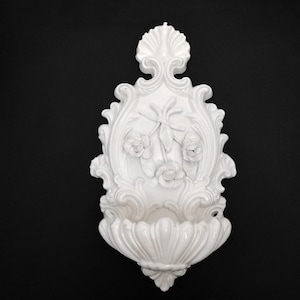 Può includere: Fontana da parete bianca e ornata con un design floreale. Presenta una cornice decorativa con dettagli a volute, tre rose e un piccolo bacino sul fondo. Lo sfondo è nero.