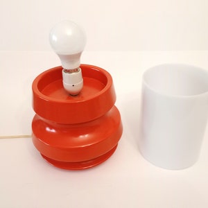 Pu&ograve; includere: Una lampada da tavolo arancione brillante con una lampadina bianca e un paralume cilindrico bianco. La lampada ha un design unico e stratificato.