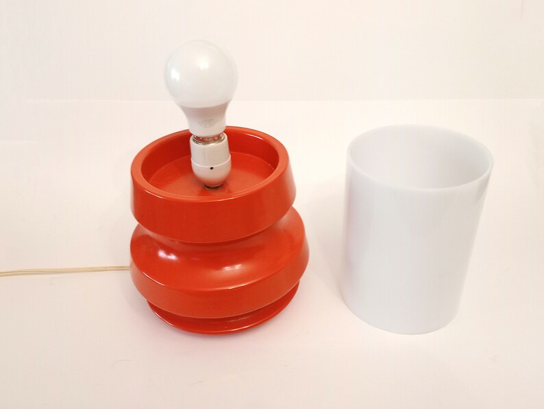 Pu&ograve; includere: Una lampada da tavolo arancione brillante con una lampadina bianca e un paralume cilindrico bianco. La lampada ha un design unico e stratificato.