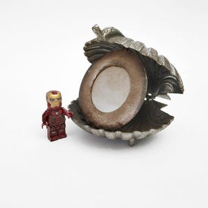 Könnte beinhalten: Eine dekorative, offene Muschelschale mit einem runden, braunen Rahmen. Eine kleine Iron Man Lego-Figur steht links von der Schale. Die Schale ist silberfarben und hat eine strukturierte Oberfläche. Der Rahmen hat eine weiße Mitte.