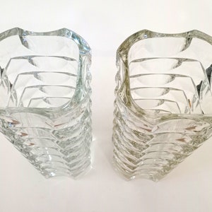 May include: Two clear glass vases with a unique, textured design. The vases have a square top that curves outward, and the sides are made up of stacked, curved sections. The clear glass allows light to pass through, creating interesting reflections.