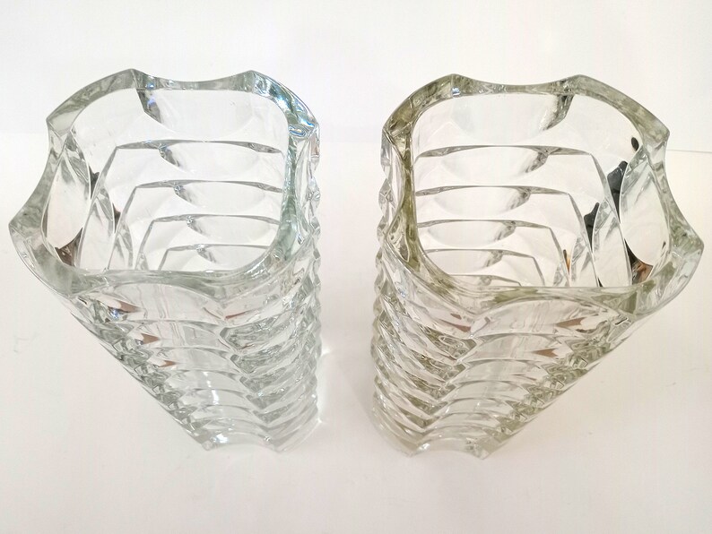 May include: Two clear glass vases with a unique, textured design. The vases have a square top that curves outward, and the sides are made up of stacked, curved sections. The clear glass allows light to pass through, creating interesting reflections.
