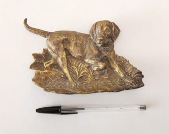 Könnte beinhalten: Eine goldfarbene Metallskulptur eines Hundes, der im Gras liegt. Der Hund ist in einer Jagdstellung, mit dem Kopf zur Seite gedreht und die Ohren aufgestellt.