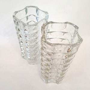 May include: Two clear glass vases with a textured, geometric design. The vases have a square shape with a wavy top edge. The glass has a faceted pattern that catches the light, creating a sparkling effect. These decorative vases are suitable for floral arrangements.