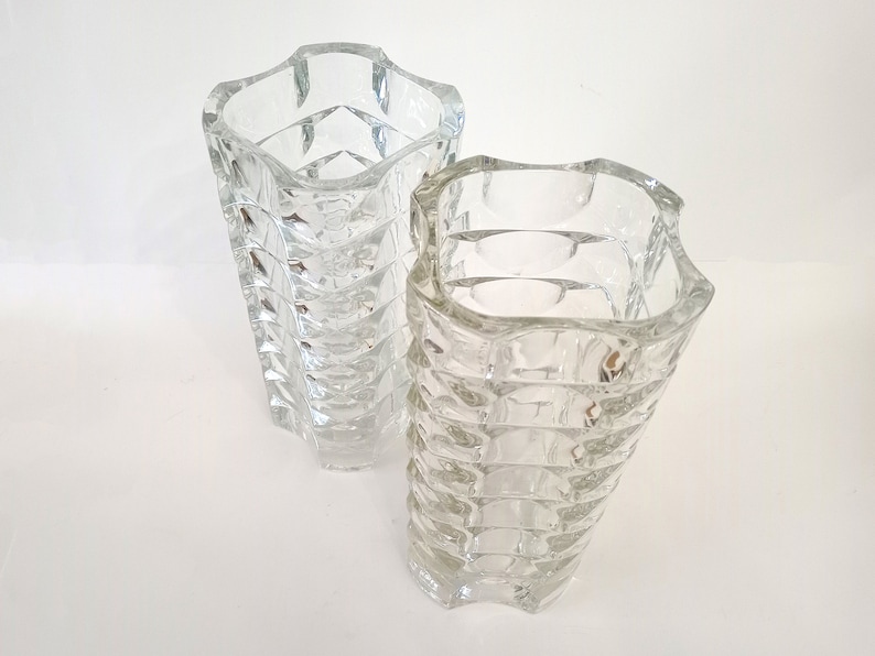 May include: Two clear glass vases with a textured, geometric design. The vases have a square shape with a wavy top edge. The glass has a faceted pattern that catches the light, creating a sparkling effect. These decorative vases are suitable for floral arrangements.