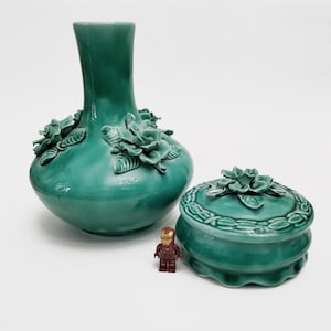 Peut inclure: Un vase en céramique turquoise et une boîte à bijoux assortie. Le vase a un corps bulbeux et un col fin, orné de roses sculptées. La boîte à bijoux est ronde avec un couvercle floral. Une petite figurine jouet est à côté des objets.