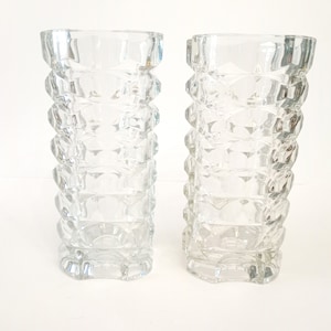 May include: Two clear glass vases with a geometric, textured design. The vases are tall and cylindrical, with a series of stacked, diamond-shaped patterns. The clear glass allows light to pass through, creating a sparkling effect. These vases are suitable for floral arrangements.