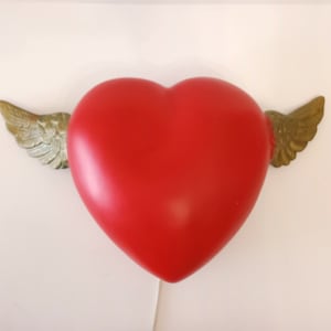 May include: A red heart-shaped ornament with bronze-coloured wings. The heart has a smooth, glossy finish, and the wings are detailed with a feathered design. The object is set against a plain white background.