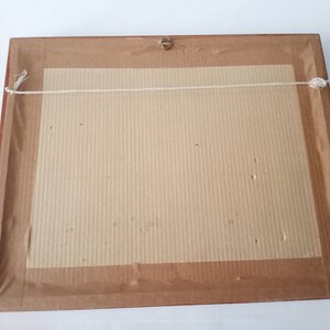 May include: The back of a framed picture with a brown wooden frame and a beige cardboard backing. The picture is hanging from a white string.