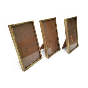 Può includere: Tre cornici rettangolari con finitura oro metallizzato e dettagli floreali decorativi agli angoli. Ogni cornice presenta un frontale in vetro trasparente e un retro marrone, posizionata verticalmente con un supporto a cavalletto.