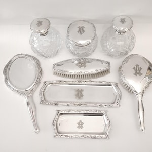 Può includere: Un set da toeletta antico con montature in metallo argentato e bottiglie di vetro trasparente. Il set include uno specchio a mano, due vassoi, due bottiglie di profumo, un barattolo e due spazzole. Ogni pezzo è decorato con un monogramma.