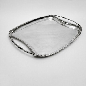 Peut inclure: Un plateau de service argenté de forme rectangulaire incurvée. Le plateau a une surface polie et des poignées avec un motif de corde torsadée. Les poignées sont intégrées au design du plateau, ajoutant un élément décoratif.