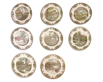May include: Set of nine vintage ceramic plates featuring countryside scenes with houses, trees, and bridges. The plates have a cream background with brown and green accents.