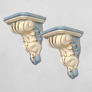 May include: A pair of decorative ceramic wall shelves. Each shelf features a light blue top and base, with a cream-coloured support adorned with gold accents and scrollwork. Designed for wall mounting, they are a decorative item.