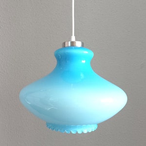 May include: A light blue glass pendant light with a rounded, bell-like shape. The glass has a gradient effect, transitioning from a darker blue at the top to a lighter blue at the bottom. The light fixture is suspended by a white cord and has a silver-colored metal cap.