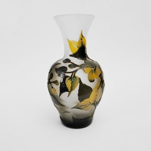 May include: A decorative glass vase with a flared top and a rounded body. The vase features a design of yellow and green leaves and flowers against a gradient background that transitions from clear to dark gray. The vase is an elegant home decor item.