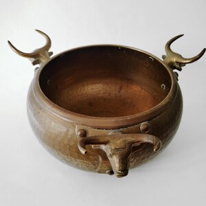 Pode incluir: Uma taça redonda de metal martelado em tom de bronze com três pegas decorativas em forma de cabeças de touro com chifres curvos. O interior da taça é liso e escuro, enquanto a superfície exterior apresenta um acabamento texturizado.