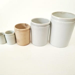 May include: Five antique stoneware crocks of varying sizes, arranged in a row. The crocks range in color from light gray to tan. The crocks are cylindrical with a slightly flared rim. The crocks are likely used for food storage.
