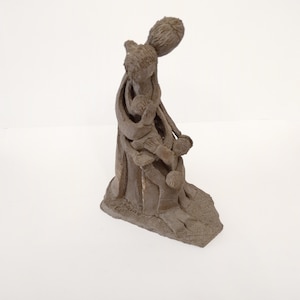 Könnte beinhalten: Eine braune Tonskulptur einer Mutter, die zwei Kinder hält. Die Skulptur ist in einer einfachen, stilisierten Art und Weise gefertigt.