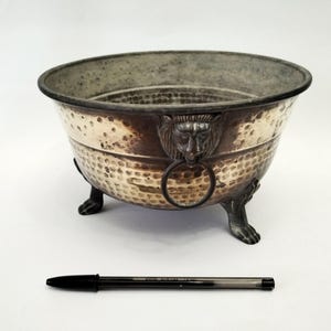 May include: A silver-toned metal bowl with a hammered texture, featuring lion head handles with rings and paw feet. The bowl has a dark rim and is set against a white background. A black pen is in the foreground.