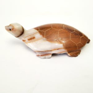 Pode incluir: Uma estatueta de tartaruga em pedra esculpida. A tartaruga tem uma carapaça castanha e branca com um padrão geométrico. A cabeça e as patas são de uma cor mais clara, e os olhos são pequenos pontos pretos. A estatueta está sobre um fundo branco.