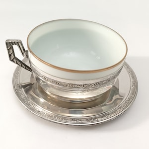 Op de afbeelding: Witte theekop met een gouden rand, op een zilveren voet en schotel. Het zilver heeft gedetailleerde geometrische patronen. Een traditionele theeset voor thee of koffie.