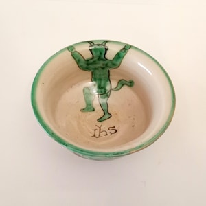 Könnte beinhalten: Eine kleine, weiße Keramikschale mit grünem Rand und einem grün bemalten Bild eines Teufels mit Hörnern und einem Schwanz. Die Schale trägt auch die Aufschrift "IHS" in Schwarz auf dem Boden.