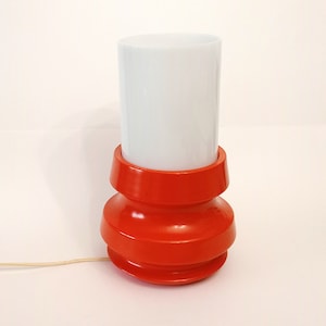 Pu&ograve; includere: Una lampada da tavolo rossa e bianca con un paralume cilindrico bianco e una base rossa con un design testurizzato.