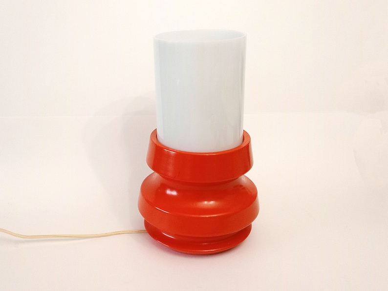 Pu&ograve; includere: Una lampada da tavolo rossa e bianca con un paralume cilindrico bianco e una base rossa con un design testurizzato.