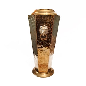 Könnte beinhalten: Eine hohe, konisch zulaufende Vase aus gehämmertem Messing mit goldfarbenem Metallic-Finish. Das Gefäß hat einen sechseckigen Korpus und ein silberfarbenes Löwenkopf-Ornament in der Mitte, das einen runden Messingring im Maul hält.