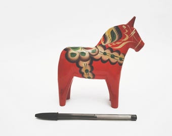 May include: A red wooden horse figurine featuring hand-painted floral patterns in green, yellow, and black along its back and mane. A black ballpoint pen is positioned horizontally in front of the horse to provide a scale reference for size.