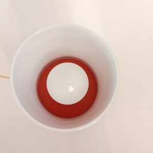 Pu&ograve; includere: Una lampada in ceramica bianca con un paralume interno rosso e una lampadina bianca. La lampada &egrave; accesa e la lampadina &egrave; illuminata.