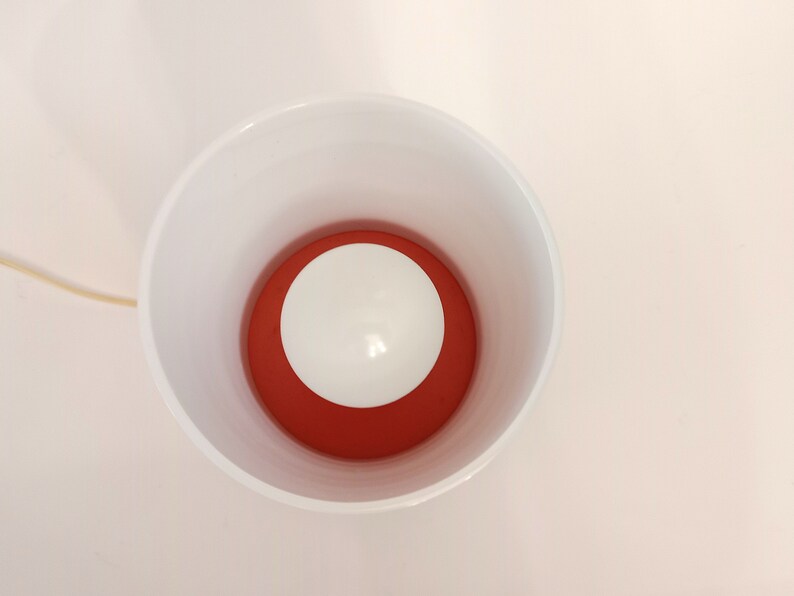 Pu&ograve; includere: Una lampada in ceramica bianca con un paralume interno rosso e una lampadina bianca. La lampada &egrave; accesa e la lampadina &egrave; illuminata.