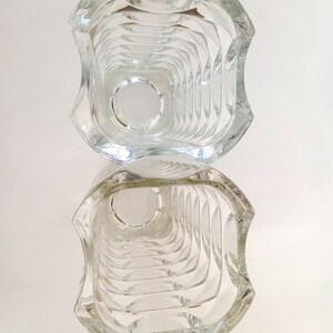 May include: Clear glass vase with a unique, geometric design. The vase has a square shape with curved edges and a textured interior. The design creates a play of light and shadow, enhancing its visual appeal. The vase is empty and reflects the light.