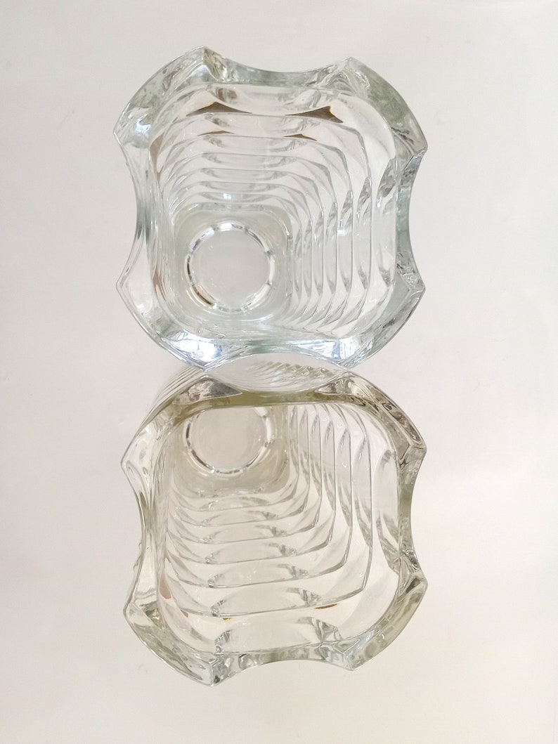 May include: Clear glass vase with a unique, geometric design. The vase has a square shape with curved edges and a textured interior. The design creates a play of light and shadow, enhancing its visual appeal. The vase is empty and reflects the light.