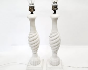 Może przedstawiać: Dwie białe marmurowe lampy stołowe ze spiralnym wzorem na kolumnie. Każda lampa ma żarówkę na górze i stoi na kwadratowej podstawie. Pióro znajduje się na pierwszym planie.