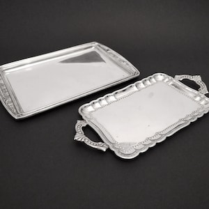 Peut inclure: Deux plateaux de service argentés. L'un est rectangulaire avec une bordure simple. L'autre a un bord festonné, des poignées ornées et une bordure perlée. Les deux plateaux sont réfléchissants et reposent sur une surface noire.