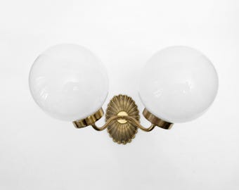 May include: A vintage wall sconce featuring two spherical, white glass globes. The fixture has a brass-colored, ornate backplate and curved arms. The design is symmetrical and suitable for interior lighting.