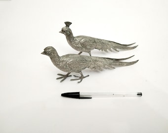 May include: Two silver-toned metal peacocks with detailed feather patterns. The peacocks are facing opposite directions and are standing on a white surface.