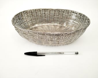 Puede incluir: Cesta ovalada de metal plateado con diseño tejido. Un bolígrafo negro y transparente se coloca debajo de la cesta. La cesta es adecuada para servir pan o exhibir objetos decorativos.