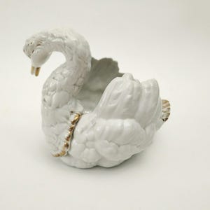 Peut inclure: Un pot de fleurs en céramique blanche en forme de cygne, avec un cou courbé et un dos ouvert. Le cygne présente des textures de plumes détaillées et des accents dorés sur le bec et autour du corps. Le pot est décoratif.