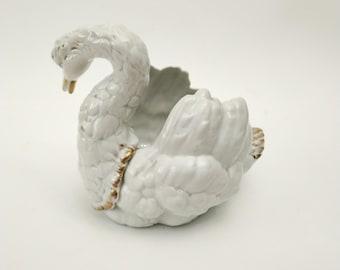 May include: A white ceramic swan-shaped planter with a curved neck and open back. The swan has detailed feather textures and gold accents on the beak and around the body. The planter is designed for decorative use.