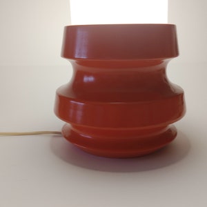 Pu&ograve; includere: Una lampada da tavolo in ceramica color terracotta con un design arrotondato e a gradini. La lampada ha un cavo e uno sfondo bianco.