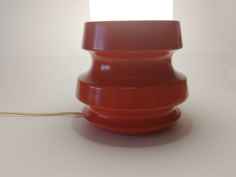 Pu&ograve; includere: Una lampada da tavolo in ceramica color terracotta con un design arrotondato e a gradini. La lampada ha un cavo e uno sfondo bianco.