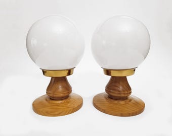 Puede incluir: Un par de lámparas de mesa con globos esféricos de cristal blanco montados sobre anillos de metal dorado. Las bases consisten en pedestales de madera torneada con un acabado natural de veta de madera en tono marrón claro.