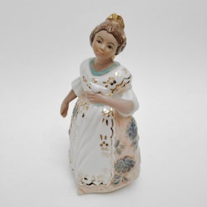 May include: A porcelain figurine of a woman in a white dress with gold accents and a blue floral pattern. The woman has brown hair and is holding a small object. The figurine is on a white background.
