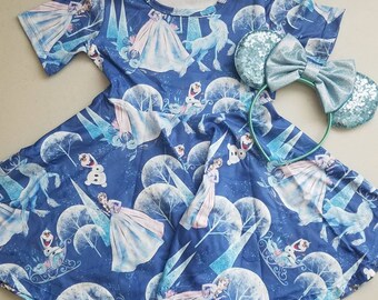 May include: A blue and white dress with a Frozen theme. The dress features images of Elsa, Anna, Olaf, and Sven. The dress has short sleeves and a flared skirt. A blue and green headband with a bow is included.